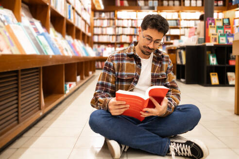 A young man sits cross-legged on the floor of a bookstore, engrossed in a red book, surrounded by shelves full of books. - ADSF54475