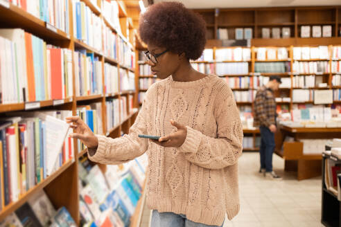 Side view of young African American woman comparing book prices on her smartphone in a bookstore - ADSF54477
