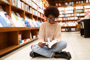 A joyful African American woman sits cross-legged on the library floor, immersed in reading a book among towering bookshelves - ADSF54478