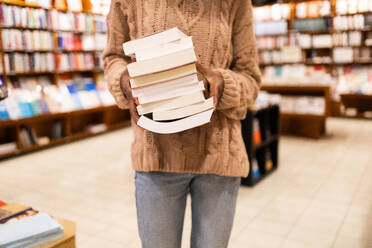 Close-up image of an unrecognizable individual in a cozy sweater holding a large pile of books, standing in the aisle of a well-stocked library - ADSF54480