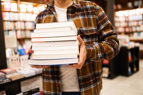 A close-up of an unrecognizable person clutching a tall stack of varied books inside a cozy bookstore, capturing the essence of literary abundance - ADSF54481