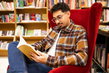 A young adult man engrossed in reading a book while sitting in a red armchair, surrounded by bookshelves in a library. - ADSF54484