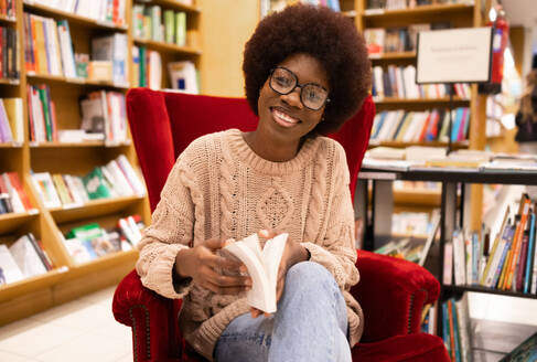 A cheerful young African American woman relaxing in a red armchair and reading a book in a cozy library setting - ADSF54485