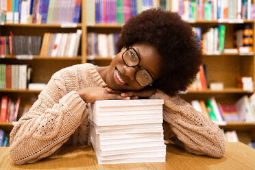 A cheerful African American woman rests her head on a stack of books in a library, surrounded by shelves of colorful literature - ADSF54487