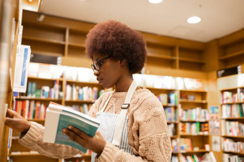 Side view of young female with an afro hairstyle is engrossed in selecting books from shelves in a cozy library setting, wearing glasses and a casual sweater - ADSF54490