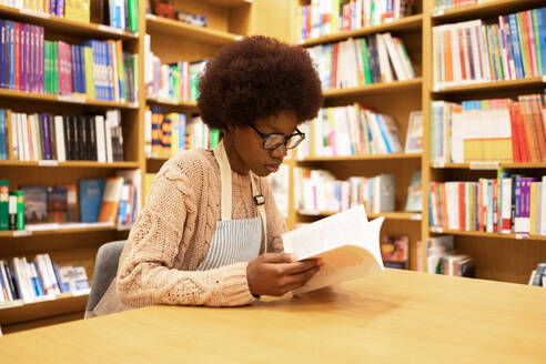 A focused young African American woman with glasses engrossed in reading a book at a well-stocked public library - ADSF54492