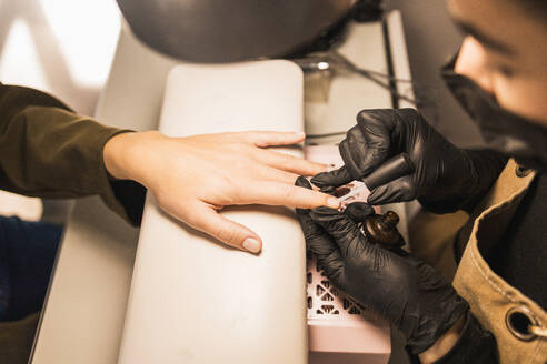 Manicurist in black gloves carefully applies brown nail polish on client’s nails during a manicure session at a beauty salon - ADSF54495