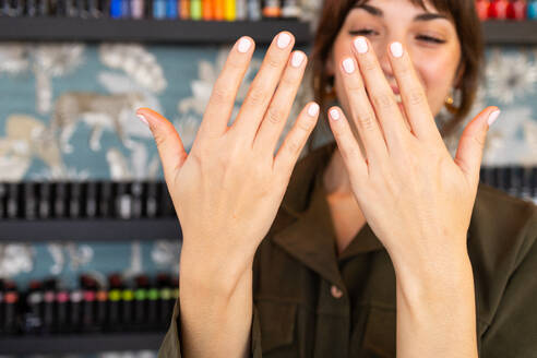 A smiling woman displays her freshly manicured nails with a subtle pink polish, in the blurred background, a variety of nail polish colors are visible - ADSF54500