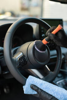 Close-up photo of hands with a brush and microfiber cloth cleaning the steering wheel in a car interior, focusing on vehicle maintenance - ADSF54522