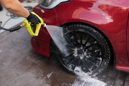 A person cleans a red car's wheel with a high-pressure washer, focusing on the rim and tire, ensuring cleanliness and maintenance - ADSF54524
