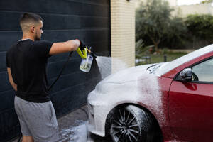 A young man is shown using a high-pressure foam sprayer to wash a red car, focusing on the cleaning process - ADSF54527