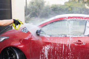 A person cleans a red car using a high-pressure washer, with white foam and water droplets visible against a blurred background - ADSF54528