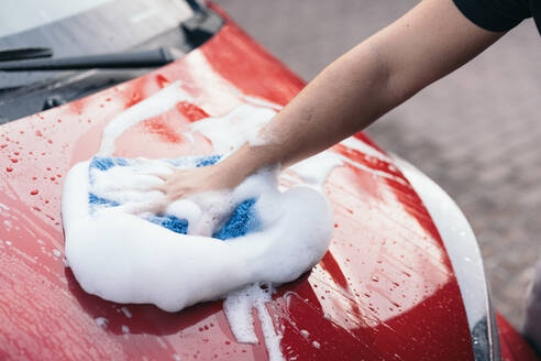 Close-up of a hand scrubbing a red car's hood with soapy foam using a blue sponge, showcasing a manual car wash - ADSF54531