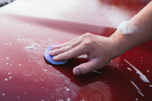 Close-up of a hand polishing a red car with a foam pad, with soap suds scattered over the surface - ADSF54532