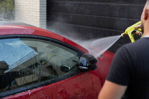 A person is using a high-pressure water spray to clean the side window of a shiny red car - ADSF54536