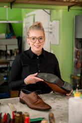 A smiling female shoemaker in Austria presents a repaired shoe at her workbench filled with tools. - ADSF54540