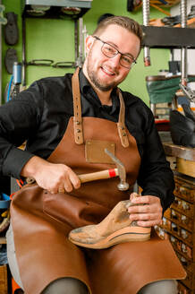 A cheerful shoemaker, wearing a leather apron, crafts a shoe with expertise in a well-equipped workshop in Austria. - ADSF54547