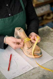 A cropped unrecognizable shoemaker in Austria meticulously measures a wooden shoe last with a yellow tape measure, surrounded by tools of the trade. - ADSF54550