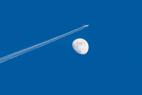 An airplane with a contrail flying past a daytime moon against a clear blue sky, captured in a tranquil and minimalist setting. - ADSF54556