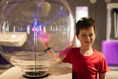A curious boy in a red shirt looks amazed at a plasma globe exhibit in a science museum, his hand interacting with the static electricity. - ADSF54575