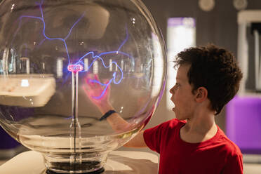 A young boy in a red shirt is fascinated by a plasma globe exhibit, touching it and observing the electrical streams at a science museum. - ADSF54577
