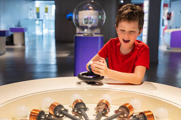Young boy in red shirt engaged with a hands-on physics exhibit, showing excitement and curiosity at a science museum. - ADSF54578