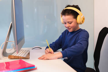 A dedicated child wearing headphones while writing notes at a desk with a computer, illustrating a modern multi-tasking study environment - ADSF54586