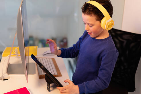 A child actively participates in an online class, gesturing while discussing or answering a question, with headphones and tablet on a desk - ADSF54587