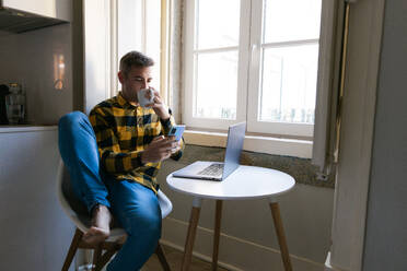 Ein entspannter Mann sitzt barfuß auf einem Stuhl und benutzt ein Smartphone, während ein Laptop und ein Kaffee auf einem kleinen Tisch neben ihm in der Nähe eines hellen Fensters stehen - ADSF54590