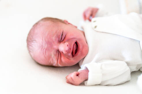 Eine Nahaufnahme, die die unverfälschte Emotion eines weinenden Neugeborenen einfängt, das in einem weichen weißen Strampler vor einem sanften Hintergrund gekleidet ist - ADSF54600