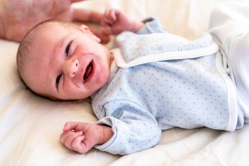 A wide-awake newborn baby in a patterned onesie lies on a soft surface, gazing with curious eyes and a partially open mouth - ADSF54602