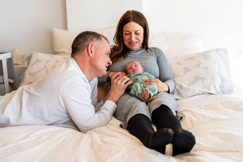 In einem gemütlichen Schlafzimmer lächeln zwei glückliche Eltern über ihr zufriedenes neugeborenes Baby, das in den Armen der Mutter ruht - ADSF54605