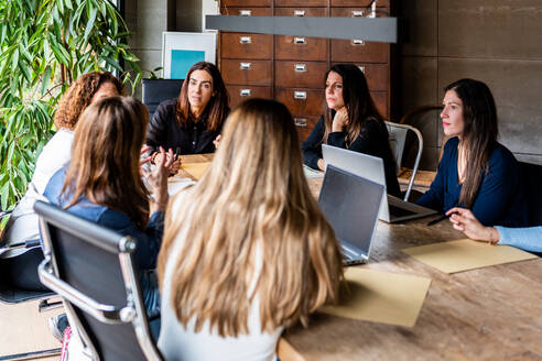 Eine Gruppe berufstätiger Frauen diskutiert an einem Holztisch in einer hellen, modernen Büroumgebung, um die weibliche Teamarbeit zu demonstrieren. - ADSF54624