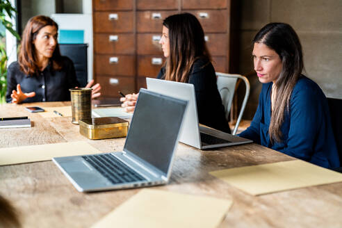 Drei Kolleginnen diskutieren an einem Holztisch mit Laptops und bringen damit die Teamarbeit in einem von Frauen geführten Unternehmen zum Ausdruck. - ADSF54629