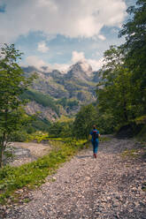 Ein einsamer Wanderer mit Rucksack wandert auf einem steinigen Pfad mitten im Grünen, mit den majestätischen französischen Alpen im Hintergrund, im Tal von Sixt-Fer-à-Cheval - ein ruhiges Ziel für Naturliebhaber und Sportbegeisterte. - ADSF54633