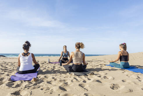 Eine Gruppe von Frauen nimmt an einer beruhigenden Yogastunde im Sand teil, mit dem Meer als Hintergrund während des Sonnenuntergangs, und verkörpert so Frieden und Achtsamkeit. - ADSF54646