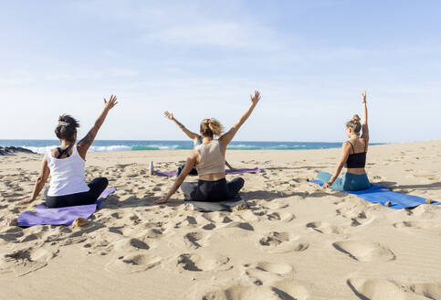 Drei Frauen praktizieren Yoga an einem Sandstrand mit dem Meer im Hintergrund während eines beruhigenden Sonnenuntergangs, wobei jede eine andere Pose einnimmt. - ADSF54652