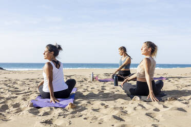 Three women practice yoga on a sandy beach with the calm ocean in the background during a tranquil sunset. - ADSF54653