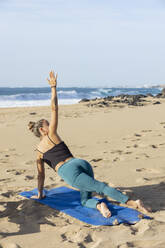 Eine Frau übt die seitliche Planke (Vasisthasana) während einer ruhigen Yogasitzung am Sandstrand vor dem Hintergrund der Meereswellen. - ADSF54657