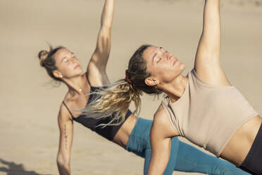 Zwei Frauen praktizieren Yoga an einem Sandstrand. Das warme Licht des Sonnenuntergangs beleuchtet ihre heiteren Gesichtsausdrücke und präzisen Posen. - ADSF54658