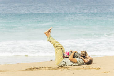 Ein fröhlicher Familienmoment, in dem ein Erwachsener und ein Kind spielerisch im Sand ringen, mit den Wellen des Ozeans im Hintergrund, als Symbol für sorglosen Strandspaß. - ADSF54680