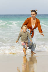 Mother and child running playfully along the beach, with waves and clear skies in the background. - ADSF54681