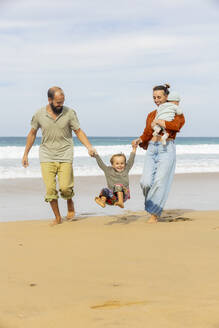 Eine glückliche Familie genießt einen spielerischen Strandspaziergang, wobei die Eltern ein entzücktes Kind an den Händen schwingen und ein Kleinkind getragen wird. - ADSF54685