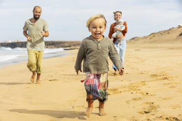Eine Familie genießt einen spielerischen Lauf am Sandstrand, mit einem fröhlichen Kind im Vordergrund, das die Essenz des Strandvergnügens einfängt. - ADSF54686