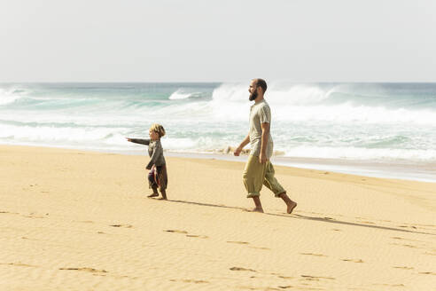 Fröhliche Familienmomente, als Vater und kleines Kind an einem Sandstrand spazieren gehen und spielen, während im Hintergrund die Wellen rauschen. - ADSF54689