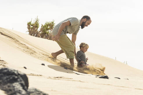 Ein kleines Kind und ein bärtiger Mann spielen in den Sanddünen unter einem strahlenden Himmel und fangen das Wesen des Familienvergnügens am Strand ein. - ADSF54690