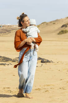 Eine fröhliche Mutter umarmt ihr Baby, während sie an einem sonnigen Strand steht und einen Moment des Familienglücks am Meer festhält. - ADSF54691