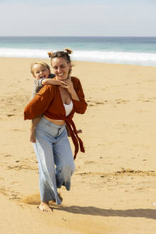 Eine lächelnde Frau nimmt ein entzücktes Kind an einem Sandstrand huckepack, mit Wellen im Hintergrund. - ADSF54693
