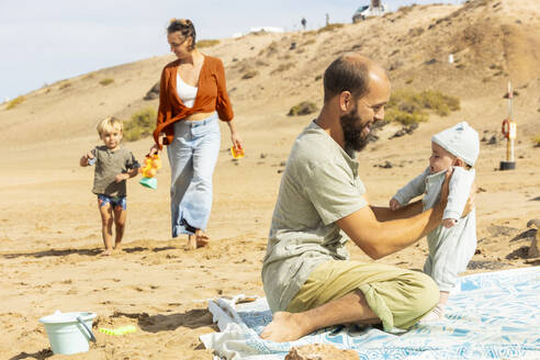 Eine Familie genießt die gemeinsame Zeit an einem Sandstrand. Der Vater spielt mit dem Baby, das Kind rennt, und die Mutter hält Spielzeug. - ADSF54695