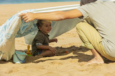 Ein Kleinkind lächelt strahlend unter einem behelfsmäßigen Zelt und genießt die Spielzeit mit einem Familienmitglied an einem Sandstrand. - ADSF54696